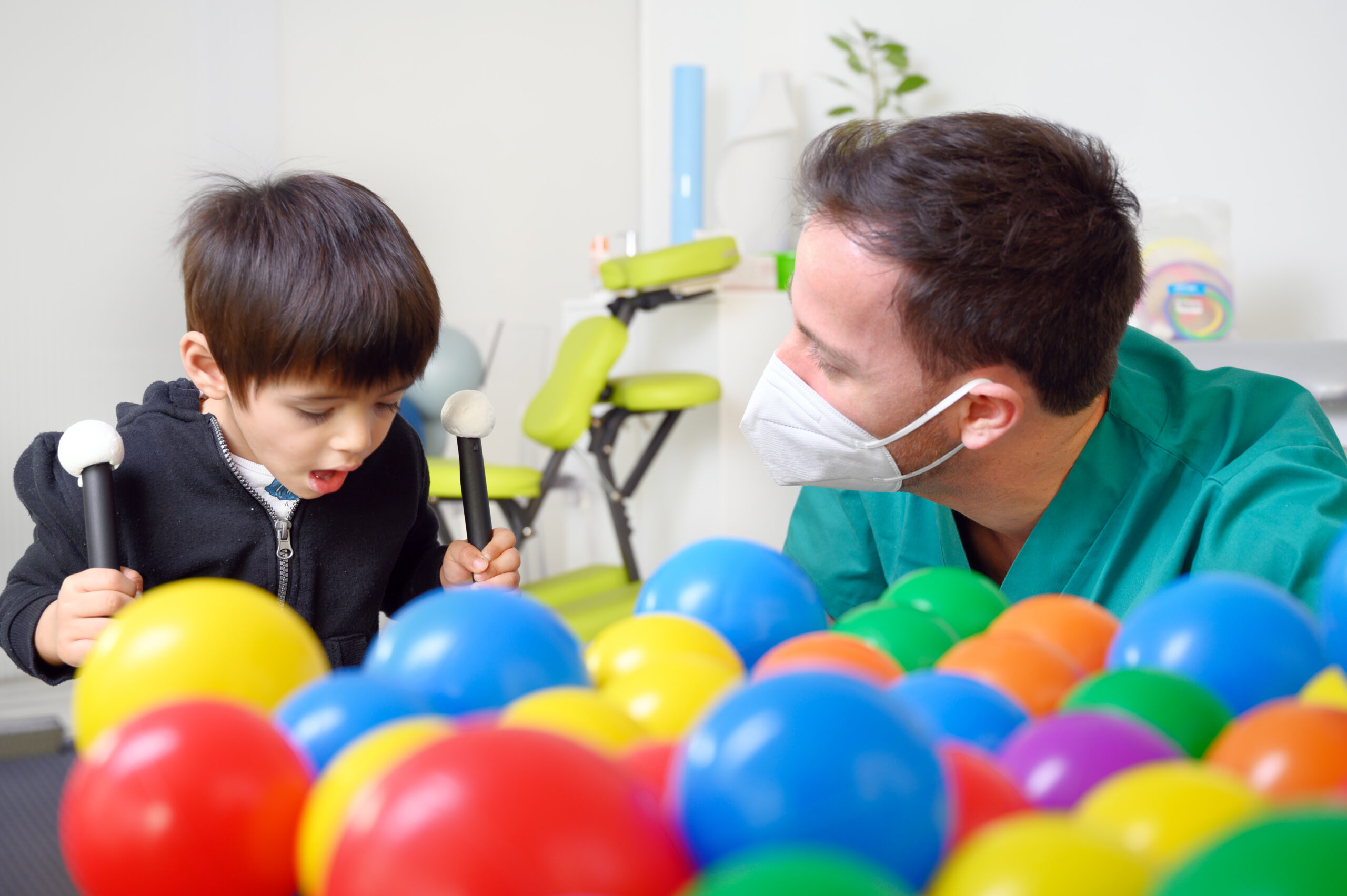 Therapist in facemask with boy with cerebral palsy behind colourful plastic balls